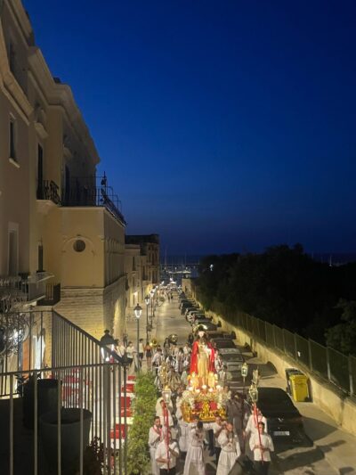 Una processione notturna si muove lungo una strada stretta in Bisceglie. I partecipanti trasportano una statua, e la scena è illuminata da lampioni.
