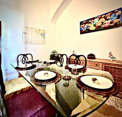 Sala da pranzo con tavolo in vetro, sedie in legno scuro e un quadro colorato appeso alla parete.